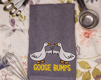 Könnte beinhalten: Ein graues Geschirrtuch mit zwei weißen Gänsen, die sich gegenüberstehen, und dem in Gelb gestickten Text "Goose Bumps". Das Handtuch liegt auf einer floral gemusterten Oberfläche mit Messlöffeln und einem Schneebesen.