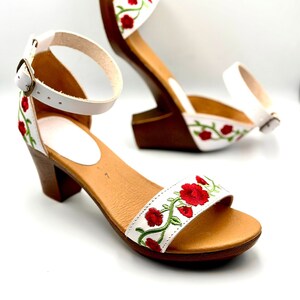 May include: White leather sandals with a red and green floral embroidery design. The sandals have a block heel and an ankle strap with a buckle closure.