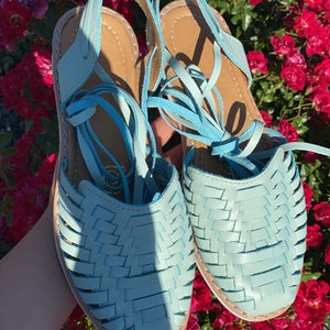 Op de afbeelding: Een paar lichtblauwe leren sandalen met geweven banden en veters. De sandalen zijn om de enkels gebonden en hebben een platte zool.