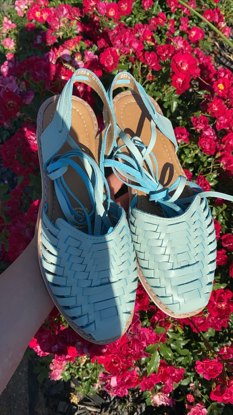 Op de afbeelding: Een paar lichtblauwe leren sandalen met geweven banden en veters. De sandalen zijn om de enkels gebonden en hebben een platte zool.