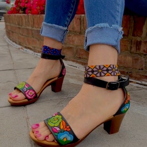 May include: Black leather sandals with a rainbow embroidered floral design and a block heel. The sandals have an ankle strap with a buckle closure. The text "Yucatan Black Rainbow" is visible below the sandals.