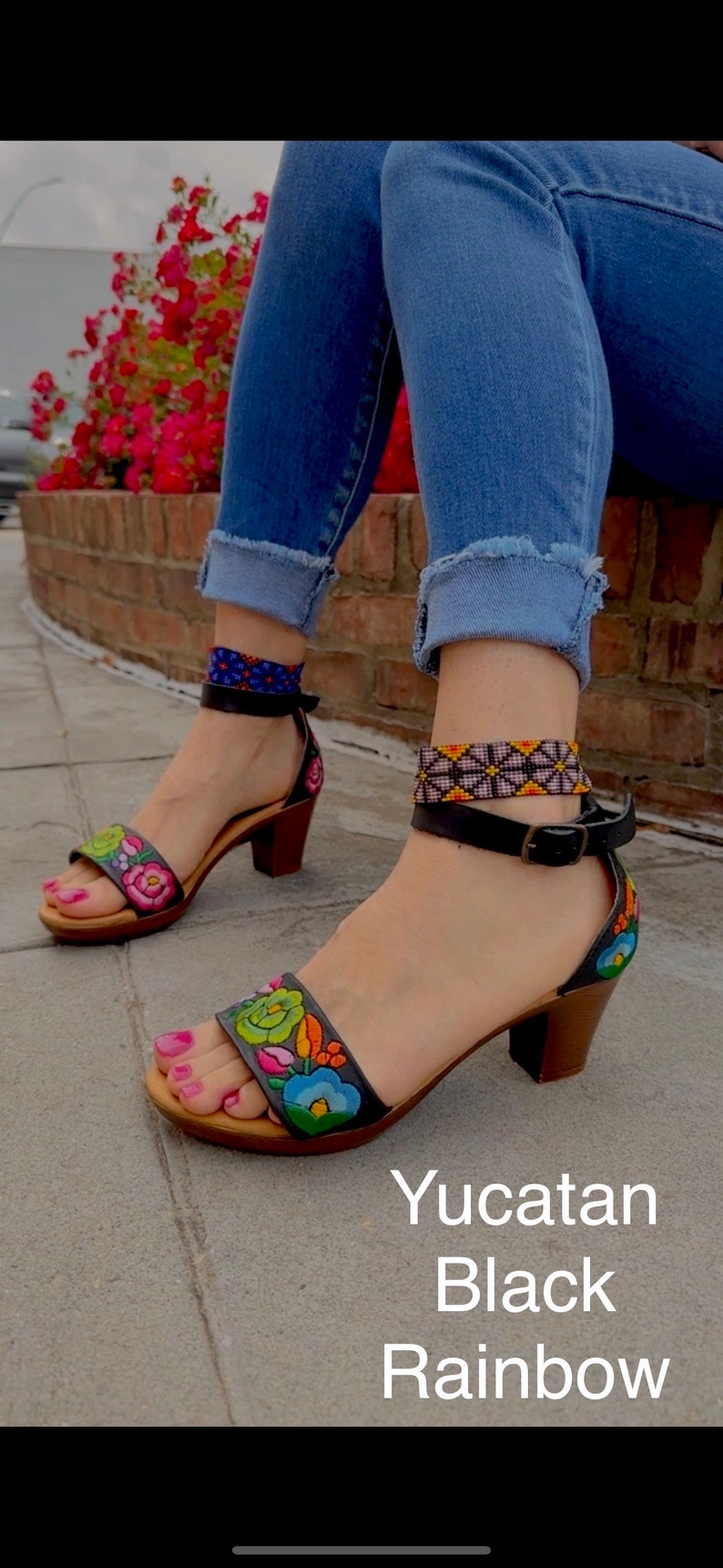 May include: Black leather sandals with a rainbow embroidered floral design and a block heel. The sandals have an ankle strap with a buckle closure. The text "Yucatan Black Rainbow" is visible below the sandals.