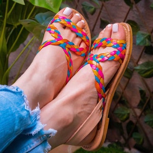 May include: A pair of sandals with braided straps in pink, blue, and yellow. The sandals are made of leather and have a tan sole.