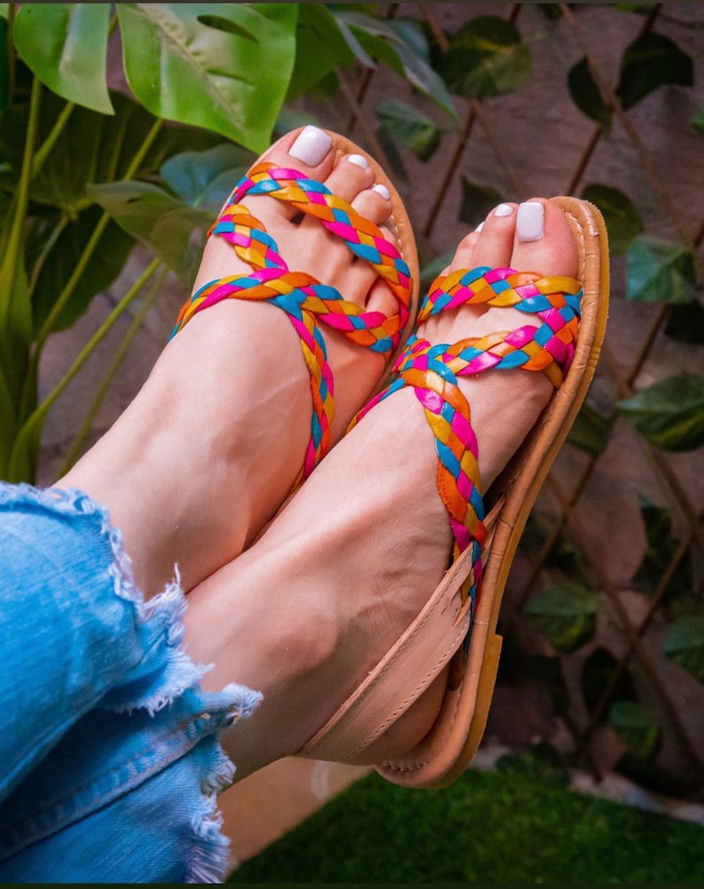 May include: A pair of sandals with braided straps in pink, blue, and yellow. The sandals are made of leather and have a tan sole.