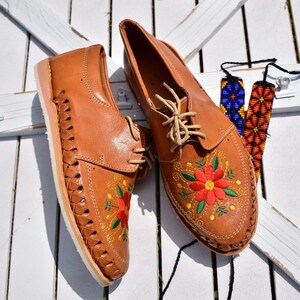 K&ouml;nnte beinhalten: Ein Paar braune Lederschuhe mit Blumenstickerei. Die Schuhe haben Schn&uuml;rsenkel und ein gewebtes Design an den Seiten. Die Schuhe befinden sich auf einer wei&szlig;en Holzoberfl&auml;che.