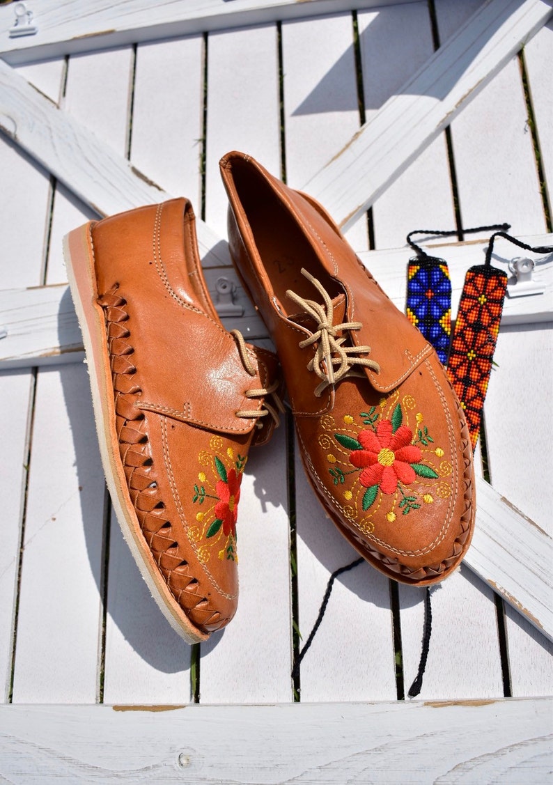K&ouml;nnte beinhalten: Ein Paar braune Lederschuhe mit Blumenstickerei. Die Schuhe haben Schn&uuml;rsenkel und ein gewebtes Design an den Seiten. Die Schuhe befinden sich auf einer wei&szlig;en Holzoberfl&auml;che.