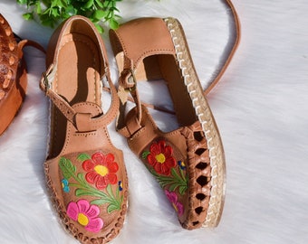 Puede incluir: Un par de sandalias de cuero marrón con bordados florales. Las sandalias tienen una suela de yute tejido y una correa para el tobillo.