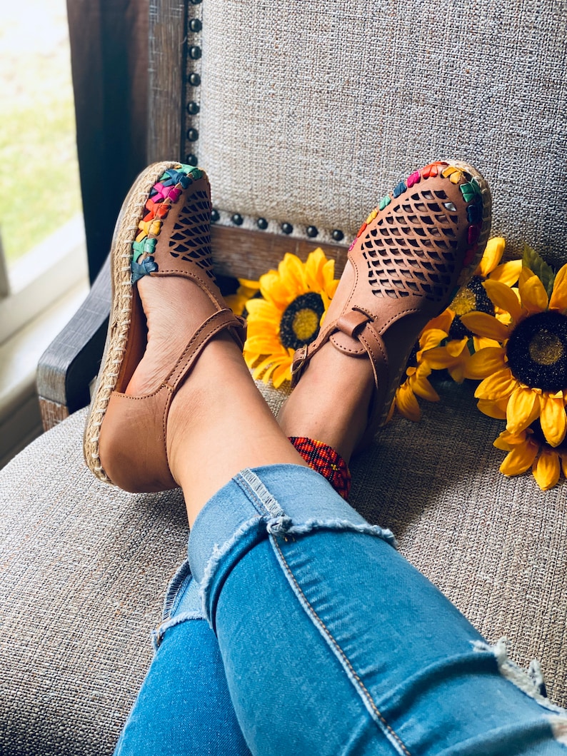 May include: Tan leather sandals with a woven colorful trim and a cut-out design. The sandals are worn by a person sitting on a chair with sunflowers in the background.