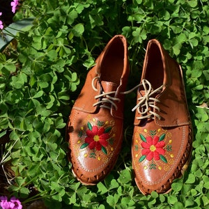 K&ouml;nnte beinhalten: Ein Paar braune Lederschuhe mit roten und gr&uuml;nen Blumenstickereien. Die Schuhe stehen auf einem Bett aus gr&uuml;nem Klee.