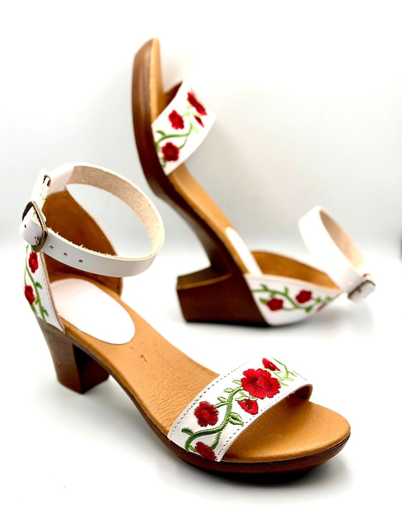 May include: White leather sandals with a red and green floral embroidery design. The sandals have a block heel and an ankle strap with a buckle closure.