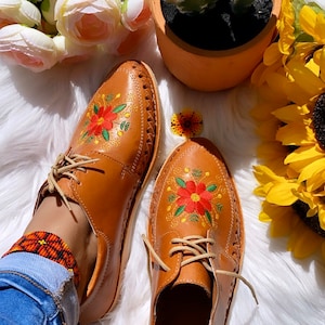 K&ouml;nnte beinhalten: Ein Paar braune Lederschuhe mit Blumenstickerei. Die Schuhe haben Schn&uuml;rsenkel und eine spitze Zehenpartie. Die Schuhe befinden sich auf einer wei&szlig;en, flauschigen Oberfl&auml;che.