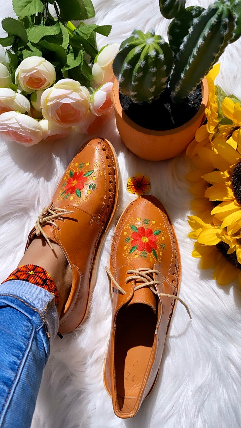 K&ouml;nnte beinhalten: Ein Paar braune Lederschuhe mit Blumenstickerei. Die Schuhe haben Schn&uuml;rsenkel und eine spitze Zehenpartie. Die Schuhe befinden sich auf einer wei&szlig;en, flauschigen Oberfl&auml;che.