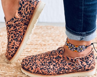 May include: A pair of brown and black leopard print woven leather sandals with white soles. The sandals are worn by a person wearing blue jeans and a beaded ankle bracelet.