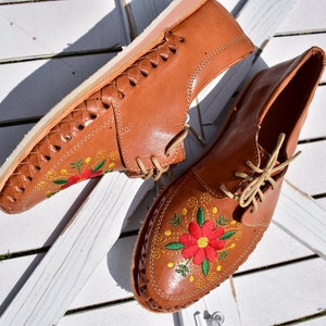 K&ouml;nnte beinhalten: Ein Paar braune Lederschuhe mit gestickten Blumen in Rot, Gelb und Gr&uuml;n. Die Schuhe haben eine geflochtene Ledersohle und Schn&uuml;rsenkel.