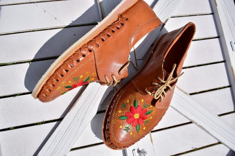 K&ouml;nnte beinhalten: Ein Paar braune Lederschuhe mit gestickten Blumen in Rot, Gelb und Gr&uuml;n. Die Schuhe haben eine geflochtene Ledersohle und Schn&uuml;rsenkel.