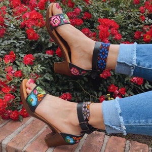 May include: A pair of black sandals with colorful embroidered flowers and a beaded ankle strap. The sandals are on a brick surface with a red rose bush in the background. The text "Yucatan Black Rainbow" is visible below the sandals.