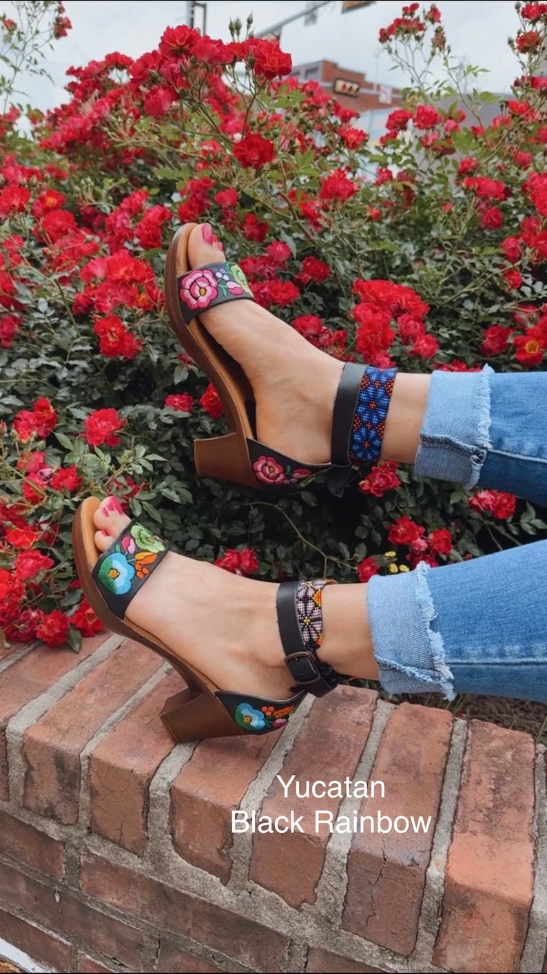 May include: A pair of black sandals with colorful embroidered flowers and a beaded ankle strap. The sandals are on a brick surface with a red rose bush in the background. The text "Yucatan Black Rainbow" is visible below the sandals.
