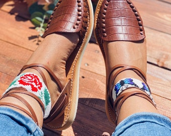 May include: Brown leather sandals with ankle straps and beaded embellishments. The sandals have a crocodile-embossed texture and are worn with blue jeans.