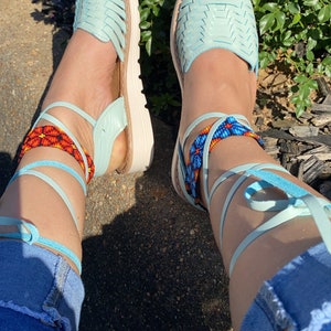 Op de afbeelding: Een paar lichtblauwe leren sandalen met geweven banden en kralen enkelbandjes. De sandalen zijn omhoog gebonden aan de benen en de persoon draagt ​​een blauwe jeans.