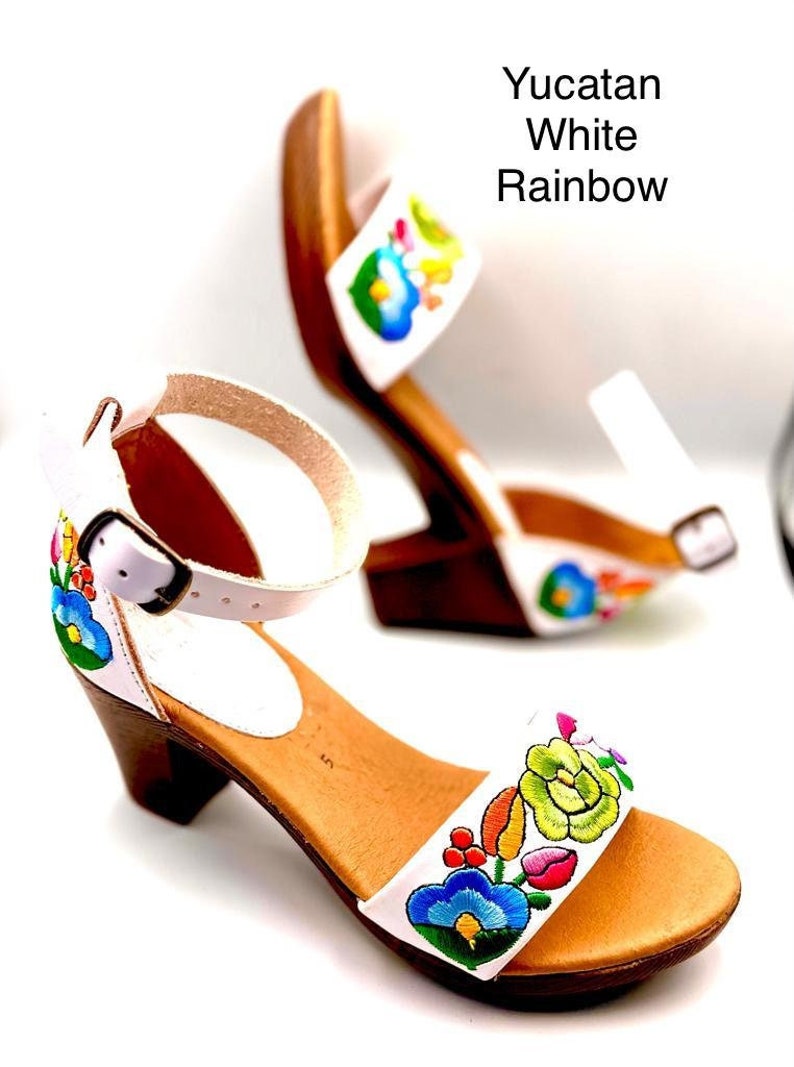May include: White leather sandals with a rainbow floral embroidery design. The sandals have a wooden heel and an ankle strap with a buckle.