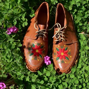 K&ouml;nnte beinhalten: Ein Paar braune Lederschuhe mit gestickten roten und gelben Blumen. Die Schuhe stehen auf einem Bett aus gr&uuml;nem Klee.