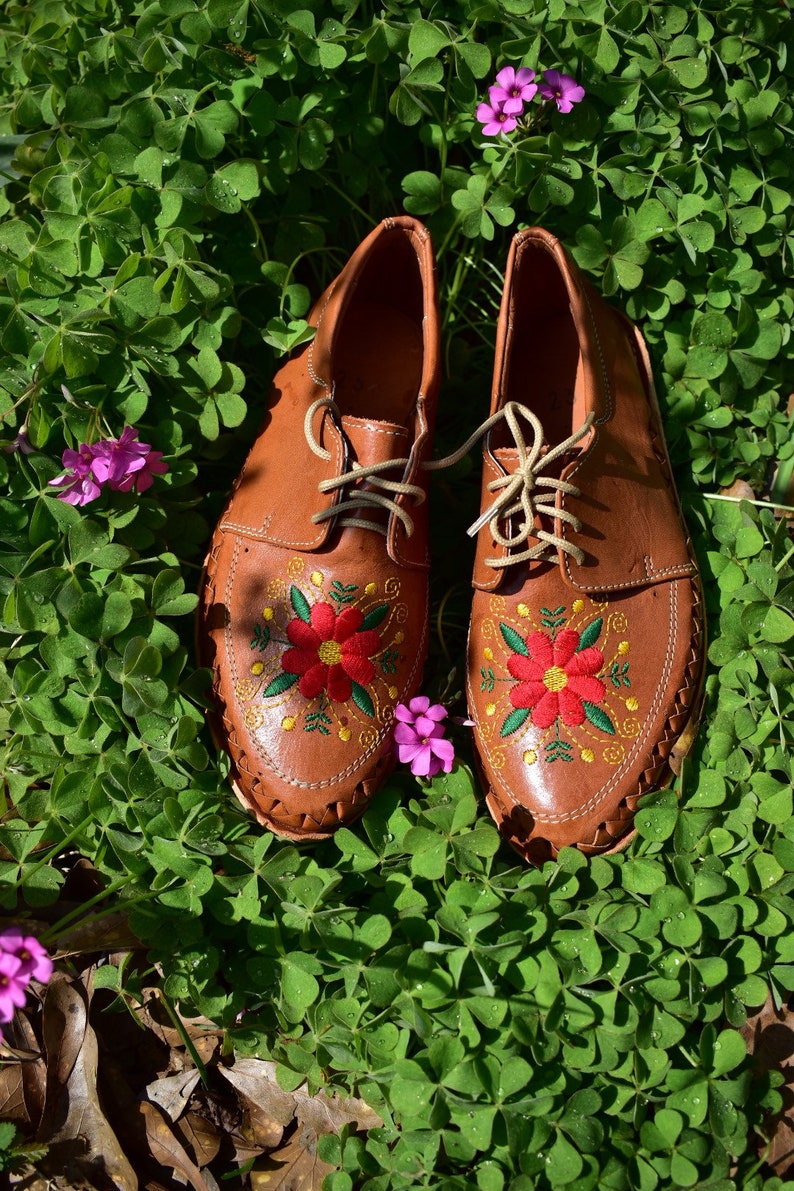 K&ouml;nnte beinhalten: Ein Paar braune Lederschuhe mit gestickten roten und gelben Blumen. Die Schuhe stehen auf einem Bett aus gr&uuml;nem Klee.