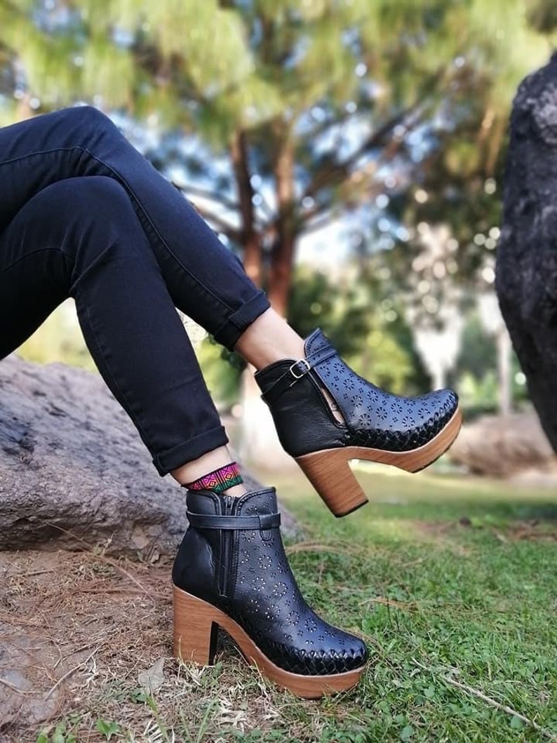 May include: Black ankle boots with a wood heel and platform. The boots have a decorative pattern of small holes and are worn with black jeans.
