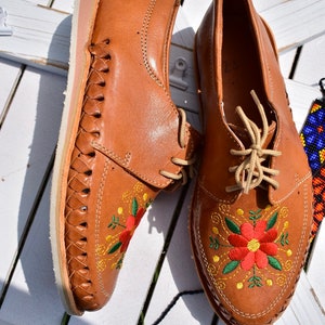 K&ouml;nnte beinhalten: Ein Paar braune Lederschuhe mit Blumenstickerei. Die Schuhe haben ein geflochtenes Lederdesign an den Seiten und eine wei&szlig;e Sohle.