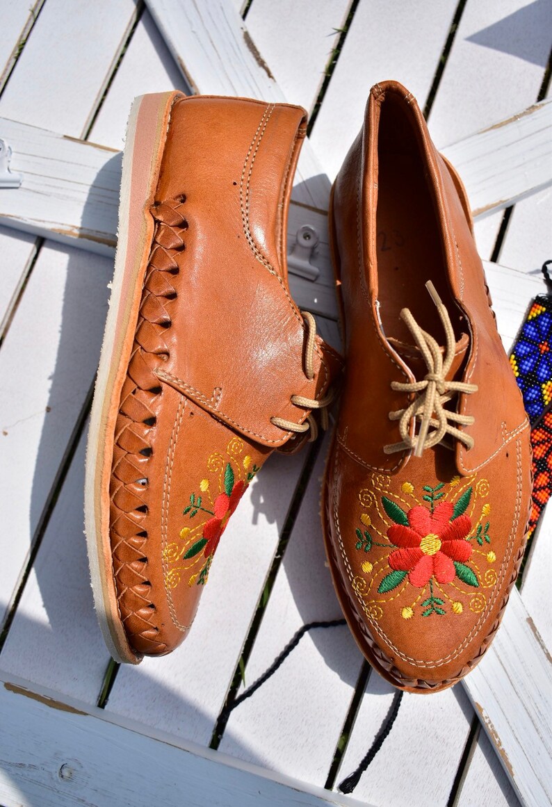 K&ouml;nnte beinhalten: Ein Paar braune Lederschuhe mit Blumenstickerei. Die Schuhe haben ein geflochtenes Lederdesign an den Seiten und eine wei&szlig;e Sohle.