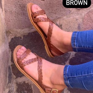 May include: A pair of brown leather sandals with a braided strap design. The sandals have a buckle closure and are worn on a person's feet.