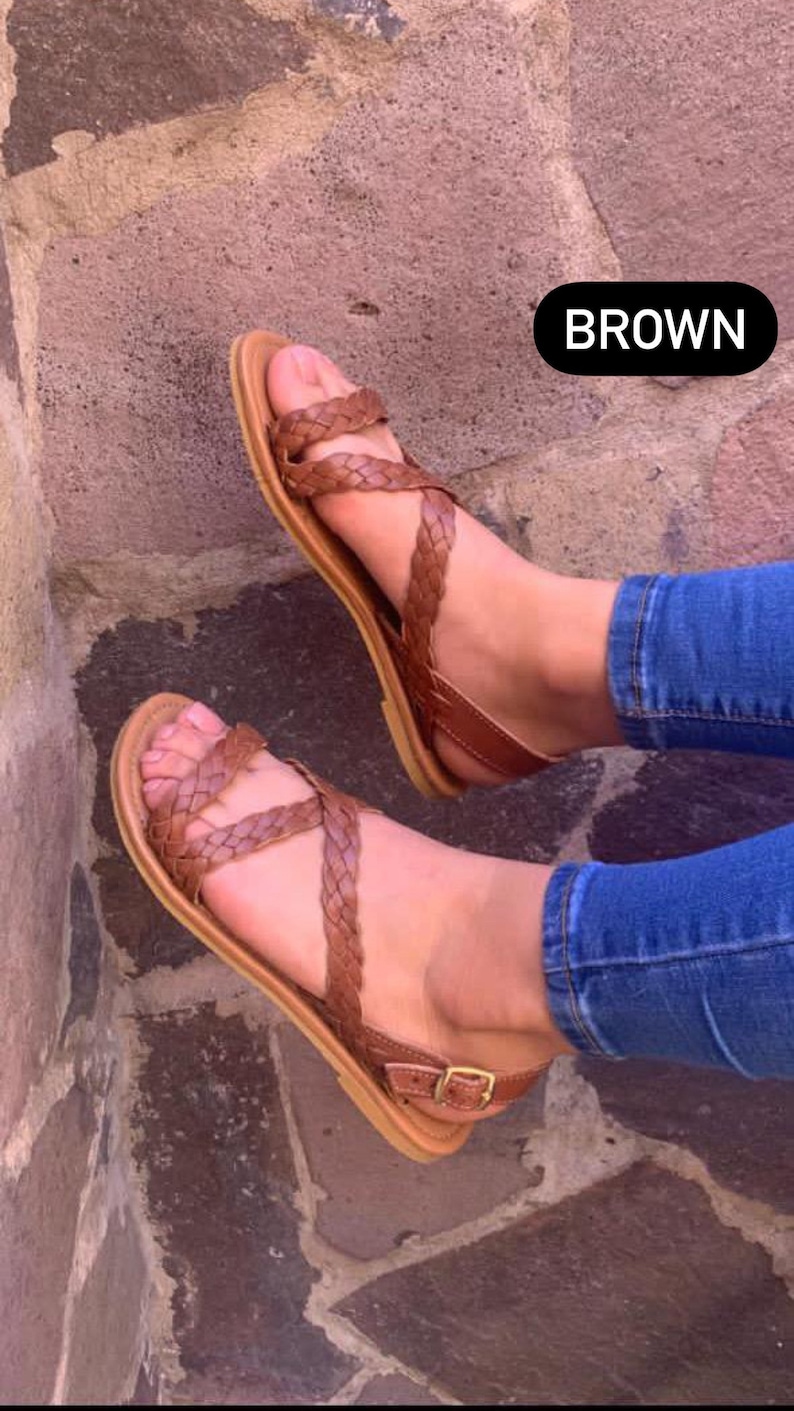May include: A pair of brown leather sandals with a braided strap design. The sandals have a buckle closure and are worn on a person's feet.