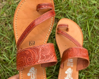 May include: A pair of brown leather sandals with intricate floral designs. The sandals have a thong style strap and a buckle closure. The sandals are made by Artesania Camilo.