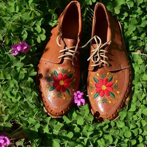 K&ouml;nnte beinhalten: Ein Paar braune Lederschuhe mit gestickten roten Blumen auf einem Bett aus gr&uuml;nem Klee.