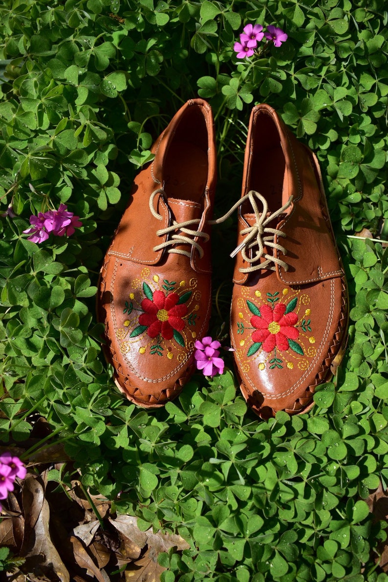 K&ouml;nnte beinhalten: Ein Paar braune Lederschuhe mit gestickten roten Blumen auf einem Bett aus gr&uuml;nem Klee.