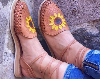 Puede incluir: Sandalias de cuero marrón con bordado de girasol y ataduras en el tobillo. Las sandalias están hechas de cuero tejido y tienen una suela plana.