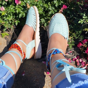 Op de afbeelding: Een paar lichtblauwe sandalen met enkelbandjes en kralenarmbanden. De sandalen worden gedragen door iemand met een blauwe jeans. De sandalen staan voor een bed van roze rozen.