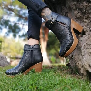 May include: Black leather ankle boots with a wood platform sole and cut-out details. The boots have a buckle closure and a zipper on the side.
