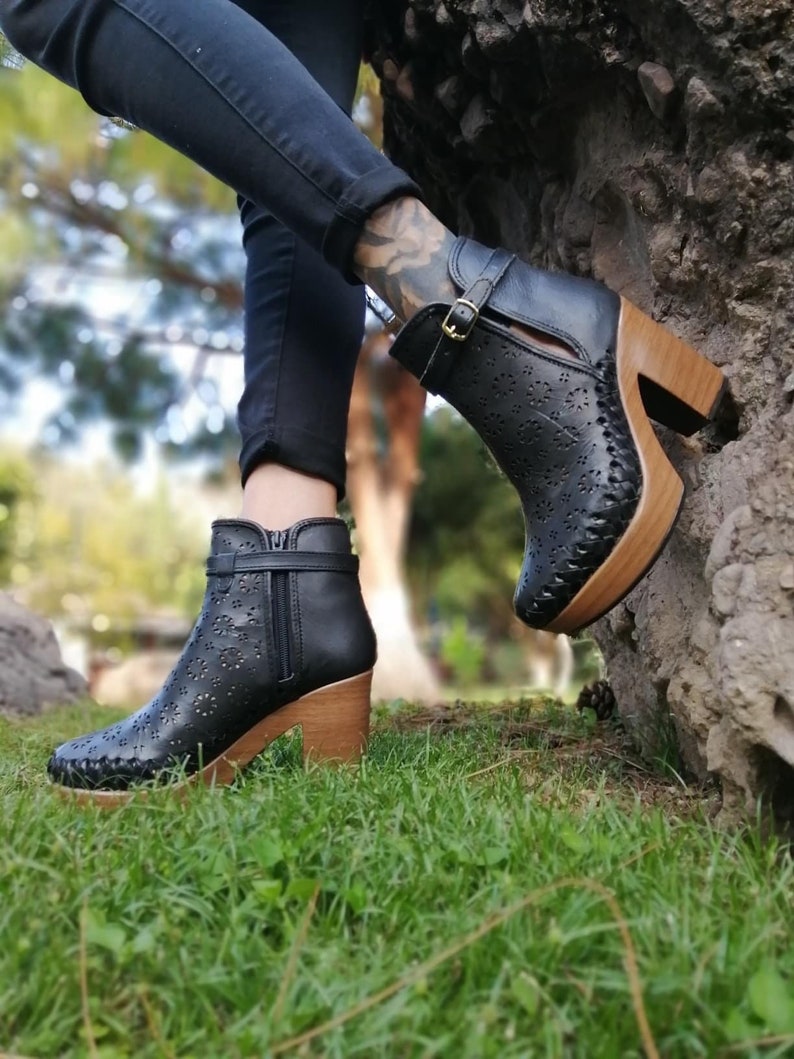 May include: Black leather ankle boots with a wood platform sole and cut-out details. The boots have a buckle closure and a zipper on the side.