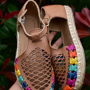 May include: A pair of brown leather sandals with a woven jute sole. The sandals have a colorful trim around the edge and a cut-out design on the upper.