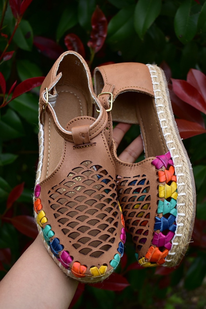 May include: A pair of brown leather sandals with a woven jute sole. The sandals have a colorful trim around the edge and a cut-out design on the upper.