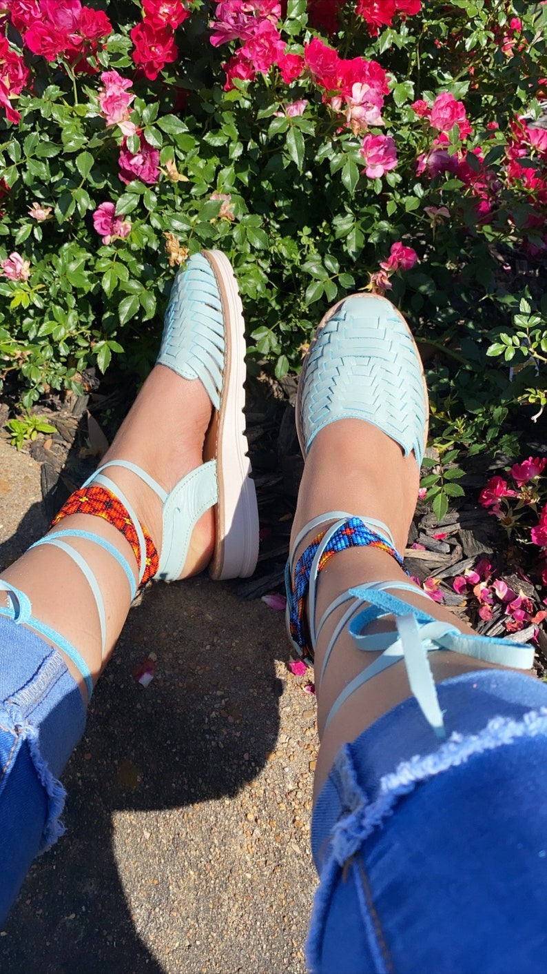 Op de afbeelding: Een paar lichtblauwe sandalen met kralen enkelbandjes. De sandalen zijn om de enkels gebonden en de bandjes zijn versierd met gekleurde kralen. De sandalen worden gedragen door iemand met een blauwe jeans.