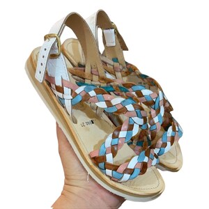May include: White leather sandals with a braided strap design in shades of brown, pink, and blue. The sandals have a buckle closure and a flat sole. The text "WHITE / RAINBOW" is visible in the image.