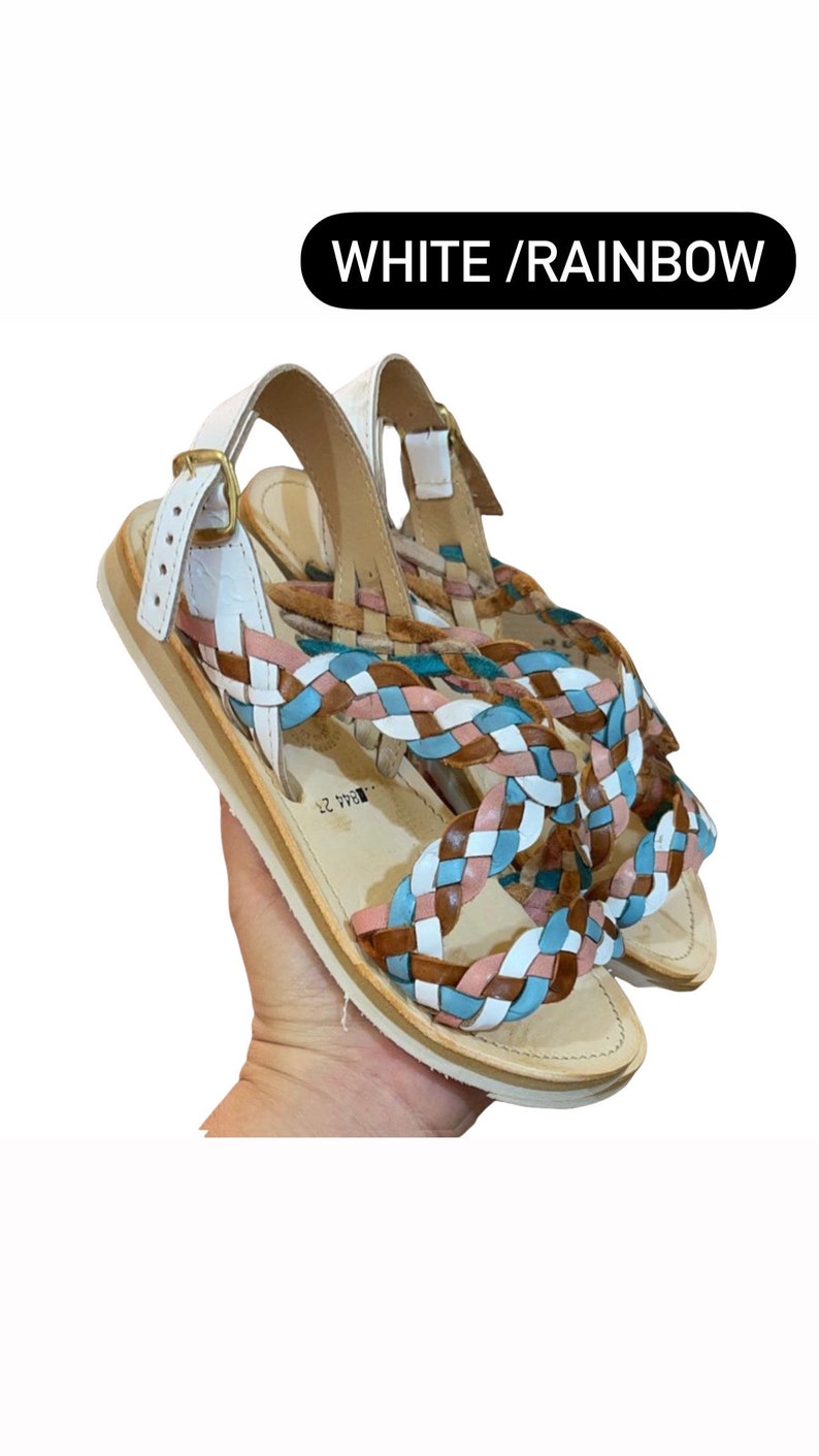May include: White leather sandals with a braided strap design in shades of brown, pink, and blue. The sandals have a buckle closure and a flat sole. The text "WHITE / RAINBOW" is visible in the image.