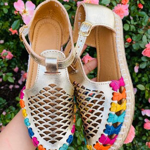 May include: Gold leather sandals with a woven rainbow trim and a cut-out design. The sandals have a buckle closure and a jute sole.