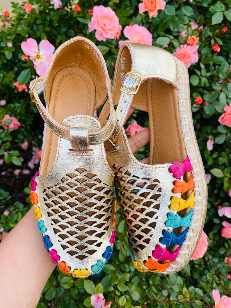 May include: Gold leather sandals with a woven rainbow trim and a cut-out design. The sandals have a buckle closure and a jute sole.