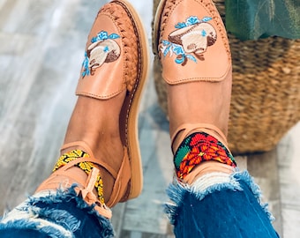 Könnte beinhalten: Ein Paar braune Ledersandalen mit gestickten Blumenmustern und einem Perlen-Knöchelriemen. Die Sandalen werden an den Füßen einer Person mit blauer Jeans getragen.