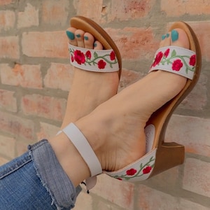 May include: White heeled sandals with a floral embroidery design featuring red flowers and green leaves. The sandals have a white ankle strap and a brown heel.