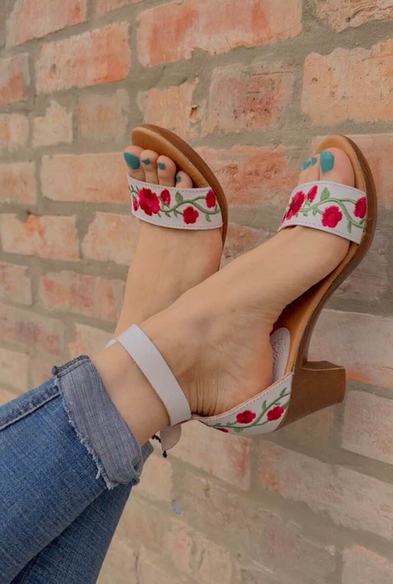 May include: White heeled sandals with a floral embroidery design featuring red flowers and green leaves. The sandals have a white ankle strap and a brown heel.