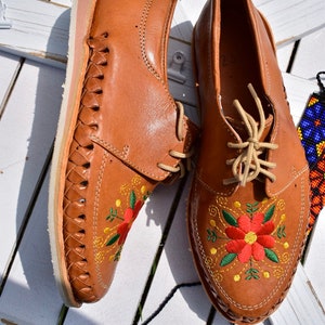 K&ouml;nnte beinhalten: Ein Paar braune Lederschuhe mit gestickten roten, gelben und gr&uuml;nen Blumen. Die Schuhe haben ein gewebtes Lederdesign an den Seiten und wei&szlig;e Sohlen.