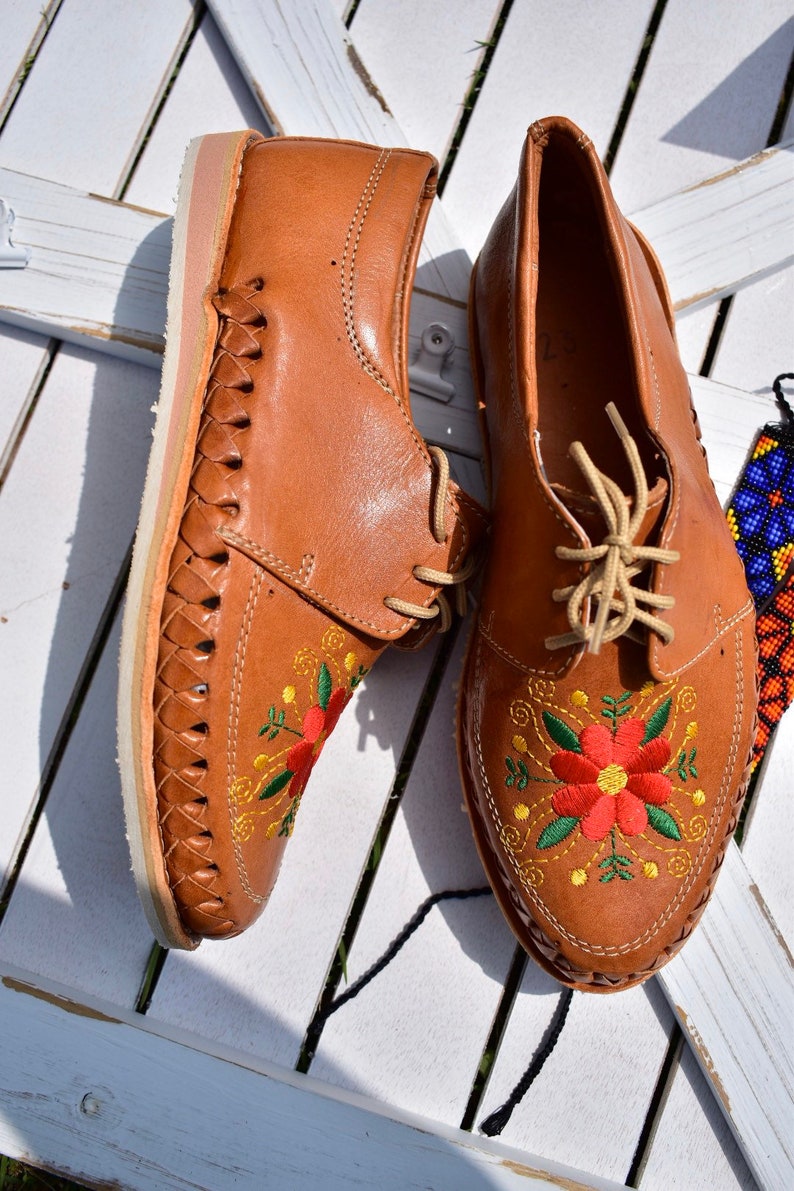 K&ouml;nnte beinhalten: Ein Paar braune Lederschuhe mit gestickten roten, gelben und gr&uuml;nen Blumen. Die Schuhe haben ein gewebtes Lederdesign an den Seiten und wei&szlig;e Sohlen.